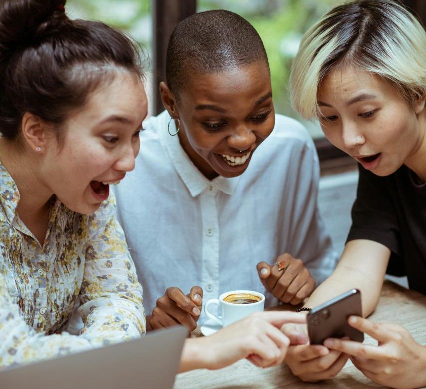 Three women sitting at a desk and looking excited at something in a phone