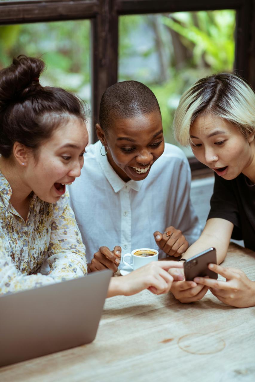 Three women sitting at a desk and looking excited at something in a phone